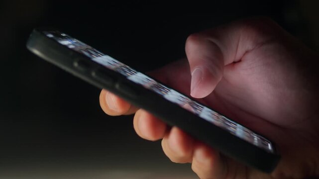 Male hand holding smartphone and scrolling social media feed in dark environment. Social media use shows modern digital lifestyle social media addiction online communication and daily scrolling habit.