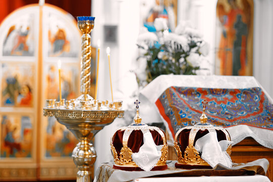 Golden Orthodox wedding crowns sit on decorated altar beside lit candles and embroidered cloth, framed by icons flowers evoke sacred tradition, reverence and celebration of church marriage ceremony