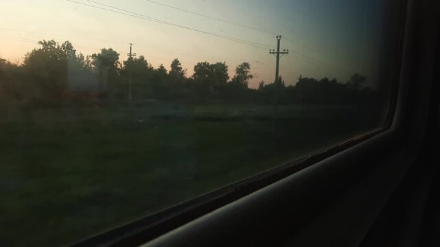A view from a moving train window onto a rural landscape at sunset with trees and an evening sky.