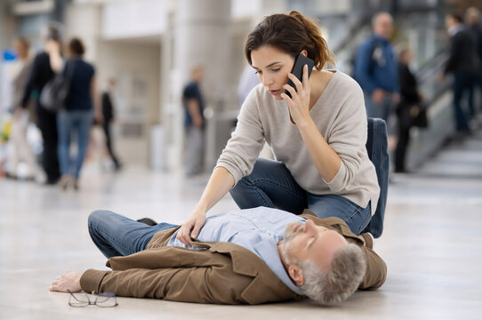 A woman calls an ambulance for a man lying unconscious on the floor in a public place. First aid is being administered. A passerby checks the condition of the fallen man.
