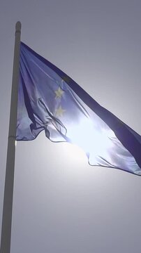 European Union flag waving majestically against a bright, sunny sky, creating a beautiful lens flare effect. The blue fabric with yellow stars ripples in the wind, symbolizing unity and prosperity