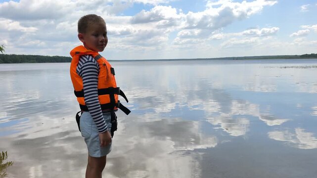 A toddler in a bright orange life jacket stands by the calm water, highlighting essential child water safety and the peace of mind that proper gear brings to family outings near lakes and rivers.