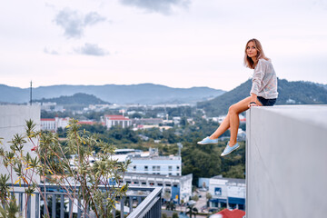 Obraz premium Young woman sits on a rooftop edge with a relaxed, contemplative expression while looking over a cityscape and distant mountains, conveying freedom, adventure, urban lifestyle and quiet reflection.