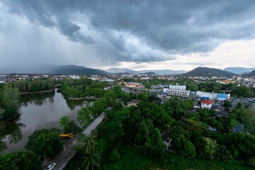 Obraz premium Aerial view of lakeside town and green suburbs under dramatic storm clouds, with distant hills, reflective water and urban buildings as heavy rain approaches, creating a moody, atmospheric landscape.