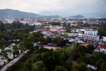 Obraz premium Tilt-shift aerial view of Phuket town with lake, trees, colorful roofs, roads and misty mountains under cloudy sky.