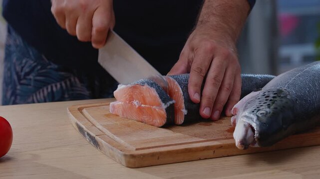 Chef cutting fresh raw salmon (trout) fish into steaks with large knife on wooden board, close-up shot.