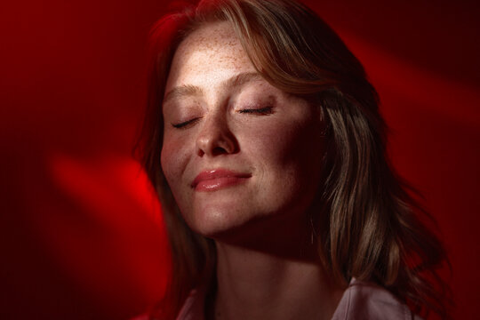 Freckled young woman with closed eyes enjoying a peaceful, relaxed moment bathed in warm red light. The intimate portrait emphasizes natural skin texture, calm expression, and soft cinematic lighting.