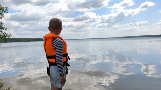 A toddler in a bright orange life jacket stands by the calm water, highlighting essential child water safety and the peace of mind that proper gear brings to family outings near lakes and rivers.