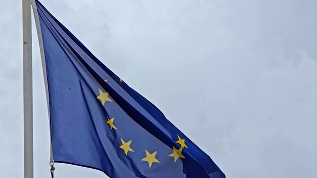 Official flag of the European Union with its circle of yellow stars waving against a grey and cloudy sky, symbolizing European unity, identity, international relations and politics