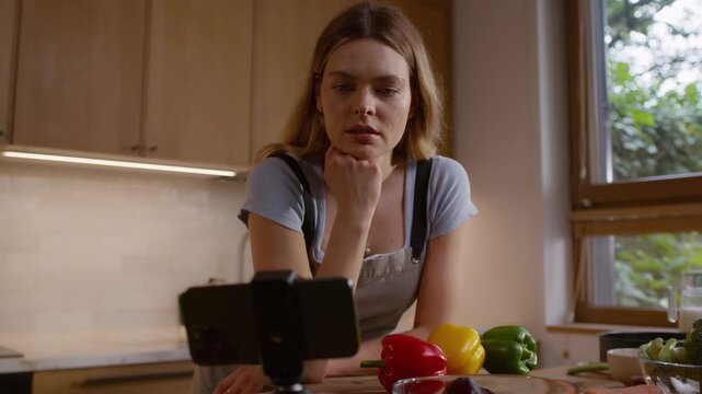 Woman leaning on the kitchen counter with a variety of vegetables broccoli and colorful bell peppers laid out on the surface. Looking into a mobile phone for recipe