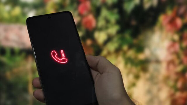 Hand holding a smartphone displaying a glowing red telephone icon with an exclamation mark, signifying an urgent call or notification against a green leafy background