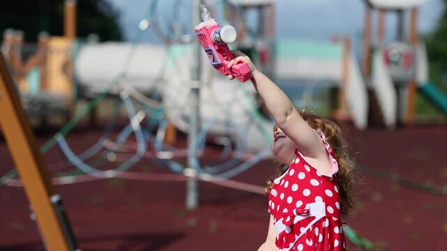 Little girl playing with bubble blower on playground. Caucasian child in red polkadot dress making soap bubbles. Happy childhood activity and outdoor summer game.