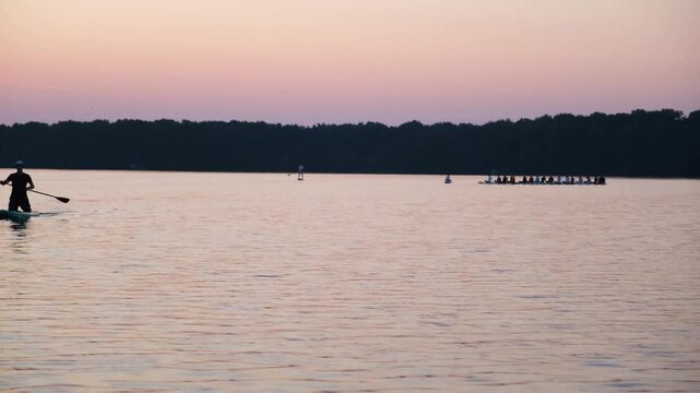 Moscow. Russia. 23.07.2026 Man standup paddleboarding on a lake at sunset. A dragon boat team rowing in the background. Summer recreational water sport activity in a calm outdoor setting.