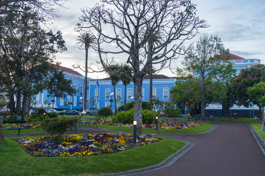 Vista de un parque o jard&iacute;n urbano con senderos, coloridos parterres de flores y &aacute;rboles, con la fachada de un elegante edificio azul tradicional en Ponta Delgada