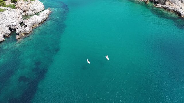 Two distant paddleboarders cross transparent coastal water above darker submerged rock formations.