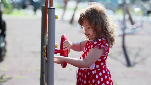 Little girl in red polka dot dress playing with steering wheel on outdoor playground. Child turning wheel toy during leisure time in sunny park summer activity.