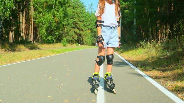 Child girl in casual clothes roller skating on road in forest on sunny summer day.