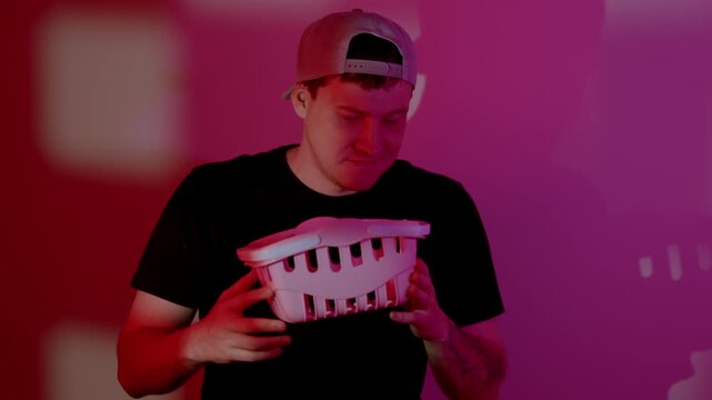Man wearing cap holds plastic basket in room with pink lighting. Young guy inspects container during routine task. Daily life activity with home equipment scene.