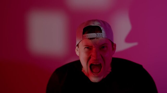Man in cap expressing rage and anger. Emotional guy shouting and grimacing with furious facial expression at camera. Aggressive reaction with intense pink lighting.