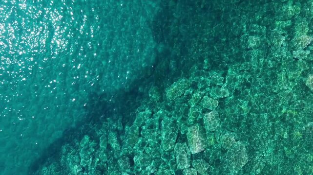 Vertical aerial view pulls back over transparent turquoise sea, underwater stones and a natural rocky coast.