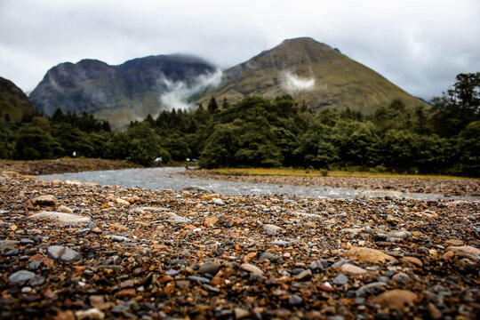 Mountains with a river lazily flowing before it in the scottish highlands. There is a small woodland, the pebbles are textured and the scene has low floating clouds. 