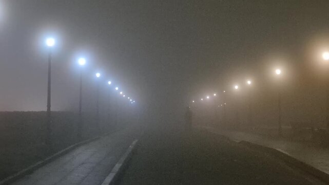 Foggy street, solitary person walking along path illuminated by glowing lamps at night in city. Static wide shot.