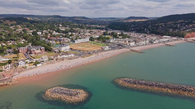 Aerial drone footage of Sidmouth in East Devon, England, showcasing the Jurassic Coast, seafront, beaches, historic town centre, and dramatic coastal scenery on a bright summer day
