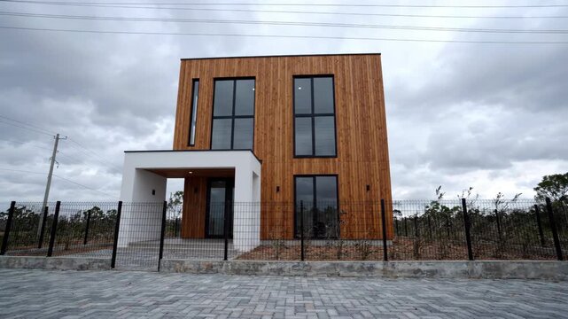 Modern Wooden House with Large Windows and Paved Driveway Under Cloudy Sky.