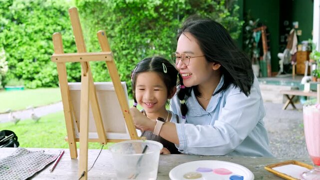 Happy Asian mother and young daughter painting on canvas together in the garden, cute little girl enjoying creative art workshop activity with mom