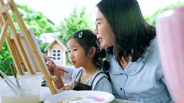 Happy Asian mother and young daughter painting on canvas together in the garden, cute little girl enjoying creative art workshop activity with mom