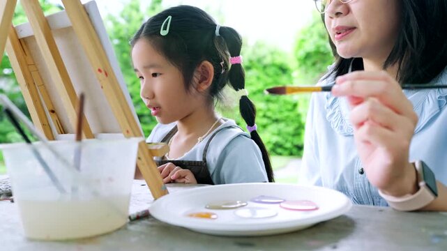 Happy Asian mother and young daughter painting on canvas together in the garden, cute little girl enjoying creative art workshop activity with mom