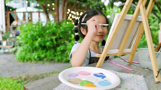 Cute happy Asian little girl showing her colorful painted hand, smiling child enjoying creative art workshop and beach canvas painting activity in the park	