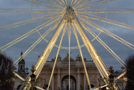 A grand Ferris wheel dominating the foreground, framing a historic, ornate stone building in the background against a dim, overcast evening sky in a city setting