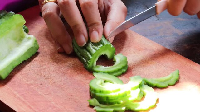 Close-up of a woman's hands slicing fresh bitter melon on a wooden cutting board while preparing a healthy homemade meal. Home cooking, vegetable preparation, and nutritious food.