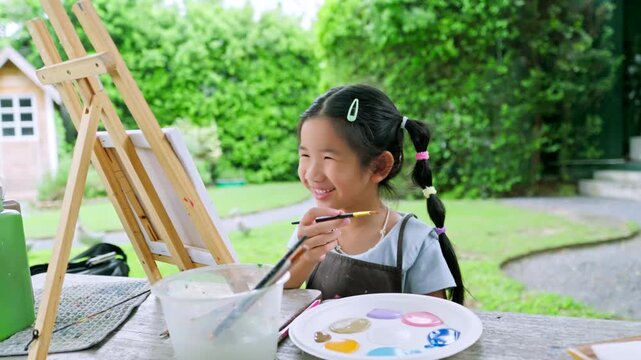 Cute happy Asian little girl showing her colorful painted hand, smiling child enjoying creative art workshop and beach canvas painting activity in the park	