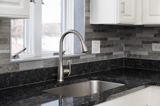 A stainless steel kitchen faucet on a black marble countertop surrounded by a grey stone tile backsplash and white cabinets.