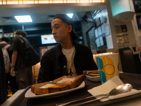 a young man wearing a backwards cap and black jacket sits in a brightly lit cafe, looking off to the side while a tray of egg toast and a paper cup sits prominently in the foreground