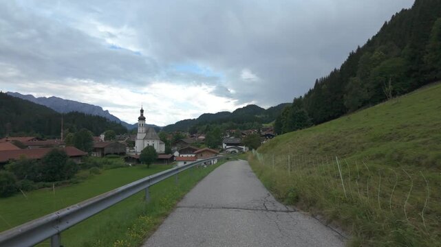 Scenic view of the alpine village of Sachrang in Bavaria surrounded by green meadows, forested hills and the beautiful German Alps under dramatic summer skies.