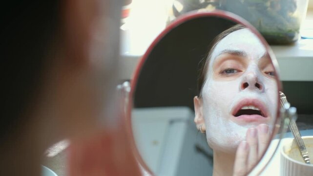 Over the shoulder view of woman looking in small mirror while applying white cosmetic clay mask to her face as part of her daily beauty routine and skincare treatment. Brunette applies anti-aging mask