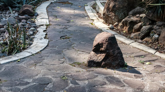 A horizontal photograph of a winding stone flagstone pathway in a botanical garden, featuring a prominent large rock in the foreground and natural landscaping with rocks and plants on the sides.