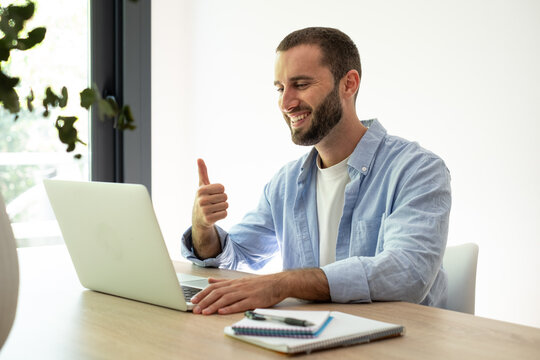 Young adult man working on a laptop at a home office, giving a thumbs up gesture, embracing innovative remote work with enthusiasm and success