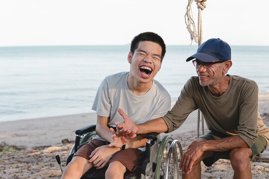 A joyful young disabled man in a wheelchair laughing out loud with his caring male caregiver sitting on a swing at the beach, showing pure happiness, family support and inclusive lifestyle.