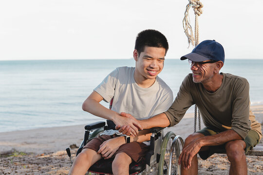 Close-up of a smiling young disabled man in a wheelchair holding hands with his caring father at the beach, showing deep trust, warm family support, love and inclusive outdoor lifestyle.