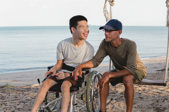 A joyful young disabled man in a wheelchair laughing while talking with his caring male caregiver on a swing at the beach, showing family support, love and inclusive outdoor lifestyle.