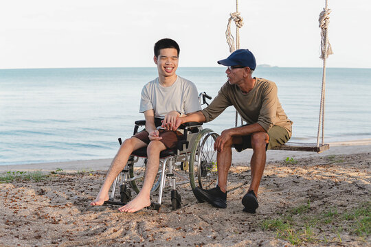 A happy young disabled man in a wheelchair smiling while holding hands with his caring father sitting on a swing at the beach, showing warm family support, love and inclusive outdoor lifestyle.