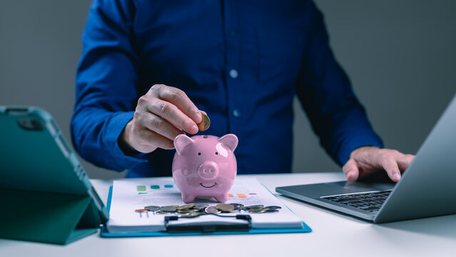 Man putting coin inside pink piggy bank sitting desk working laptop computer analyzing financial chart document planning budget saving investment strategy personal finance home office table.