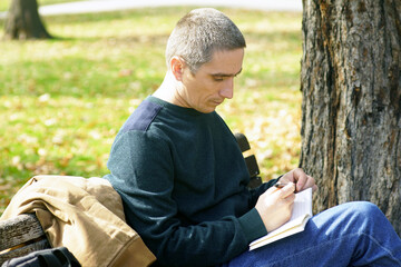 Obraz premium Man writing in notebook while relaxing beneath tree in park during calm autumn day. Represents offline reflection, mindful journaling, cognitive reset, and sustainable attention management habits.