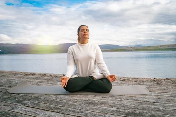 Fotobehang Lotusbloem Woman meditating in lotus positionon a wooden pier by a scenic fjord in Norway  © Muñe