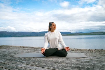 Fotobehang Lotusbloem Woman meditating in lotus position on a wooden pier by a scenic fjord in Norway  © Muñe