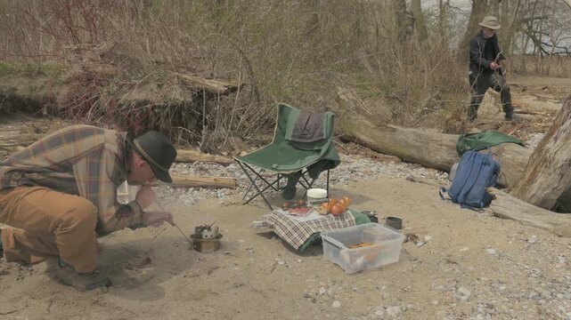 Two senior men gather fuel and tend fire in compact camp grill at lakeshore picnic site, Lynde Shores Conservation Area, Whitby, Ontario, Canada.